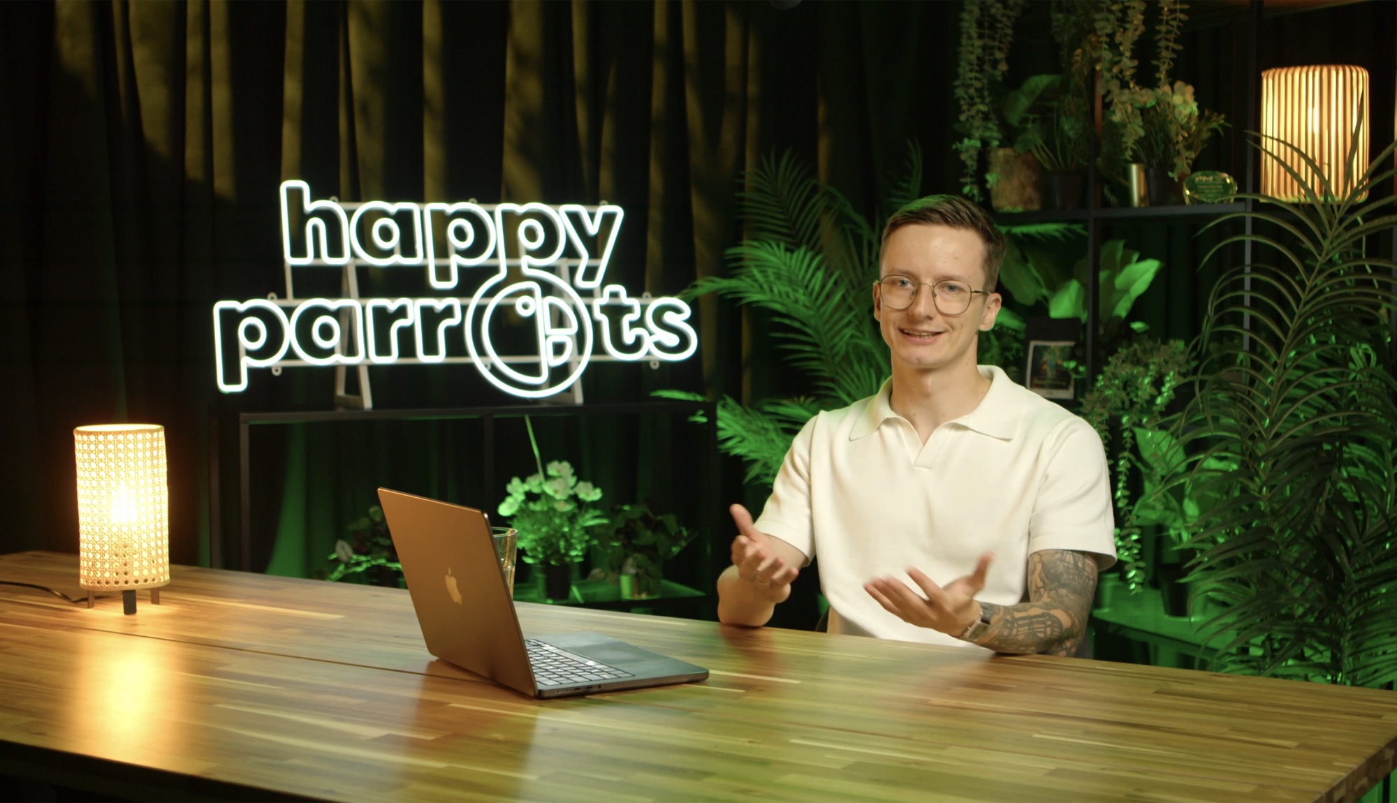 Jakub Papuga nagrywający podcast Happy Parrots, przy stole, w tle neon "happy parrots" i rośliny